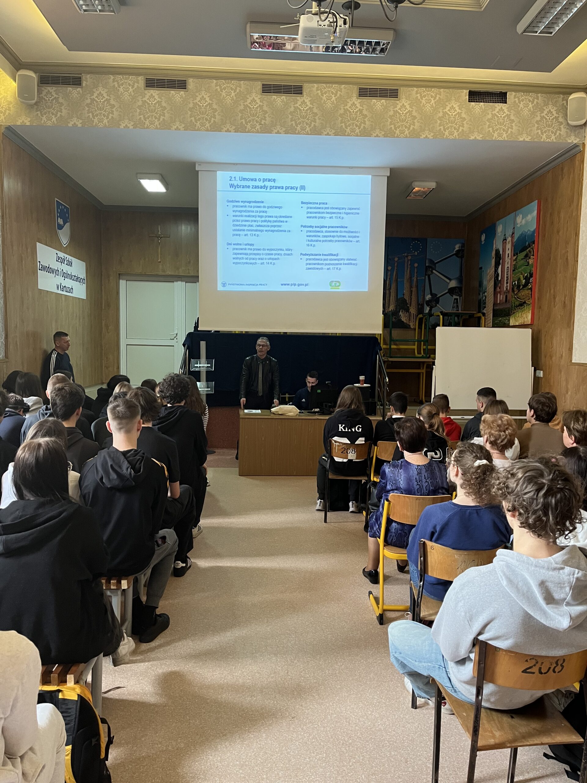 Prowadzący zajęcia edukacyjne. Uczeń siedzący przy biurku, który przesuwa prezentację pomocną przy prowadzeniu zajęć przez inspektora PIP.